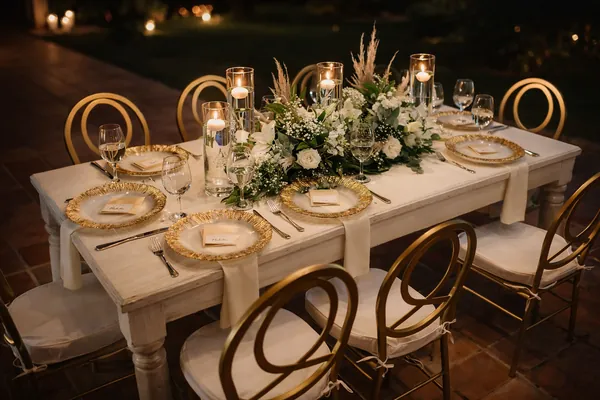 Mesa de noche con centro alargado de rosas blancas, follaje y velas, y sillas doradas