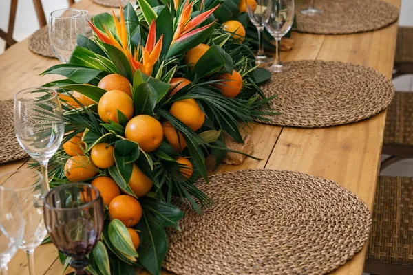 Centro de mesa alargado de aves del paraíso, naranjas y follaje sobre una mesa de madera