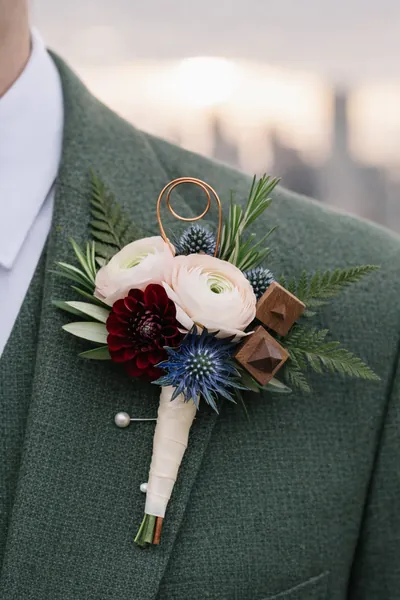 Boutonnière de ranúnculos melocotón, dalia vino tinto y cardo azul en un traje verde